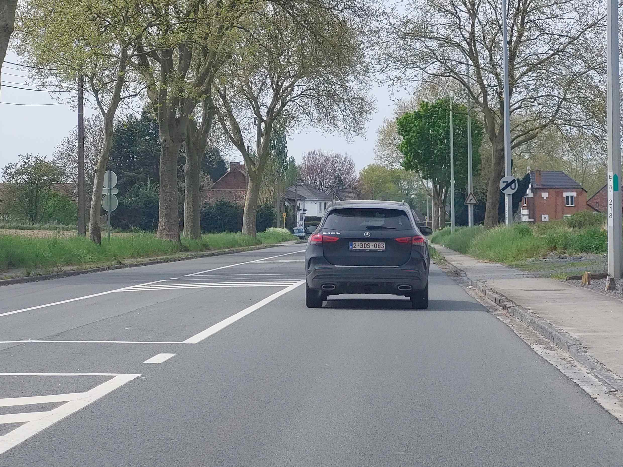 En tant qu'expert en code de la route belge, voici une description détaillée de la situation routière présentée dans l'image, en relation avec la question posée :
Contexte de la question: "Wallonie, à cet endroit, ma vitesse est limitée à :"
Description Détaillée de la Situation Routière
Type de situation routière:
L'image représente une portion de route bidirectionnelle, vraisemblablement en dehors d'une agglomération ou en bordure de celle-ci (zone semi-rurale/semi-urbaine), située en Wallonie. La chaussée principale, en direction de la caméra, comporte une voie de circulation avec un marquage au sol indiquant la préparation à une intersection ou un carrefour. Sur la droite, un accotement ou bas-côté est présent, bordé d'un trottoir et de végétation.
Éléments visibles:
Véhicules: Un véhicule de type SUV foncé (une Mercedes GLA 250, comme indiqué sur le coffre, avec l'immatriculation belge 2-BDS-083) est visible devant le véhicule de la caméra, circulant dans la même direction et occupant la voie de droite.
Panneaux de signalisation:
Sur le côté droit de la route, derrière le SUV et à hauteur du véhicule de la caméra, un panneau de signalisation circulaire à fond blanc, bordure rouge et chiffre noir "70" (panneau C43) est clairement visible. Ce panneau indique une limitation de vitesse.
Juste derrière ce panneau C43, un panneau de danger triangulaire à fond blanc, bordure noire et symbole noir représentant un virage dangereux à droite (panneau A14) est également visible, partiellement masqué par le panneau C43.
Plus loin sur la droite, un poteau de signalisation vertical porte une inscription "E181 218", qui est une borne kilométrique ou hectométrique indiquant probablement la route (E181) et le point kilométrique.
Feux de signalisation: Aucun feu de signalisation n'est visible.
Marquages au sol:
Une ligne blanche continue délimite la bande de roulement du côté droit (accotement).
Une ligne blanche discontinue sépare la voie de circulation du véhicule de la caméra d'une voie adjacente (probablement une voie de présélection pour tourner à gauche, comme suggéré par les flèches et marquages en damier sur la gauche de la photo, juste devant la position de la caméra).
Le véhicule de la caméra se trouve dans la voie de droite, tandis que des marquages au sol plus larges et des chevrons indiquent une zone de préparation à un tourne-à-gauche ou une voie de décélération/stockage pour un tourne-à-gauche.
Environnement: La route est bordée d'arbres hauts des deux côtés, dont certains sont en pleine feuillage printanière. De la végétation basse et de l'herbe s'étendent le long de la route. Plusieurs habitations de type maisons individuelles en briques sont visibles en arrière-plan, suggérant une présence résidentielle à proximité. Un trottoir en bordure de la route est visible sur la droite.
Position et direction des véhicules:
Le véhicule de la caméra est en approche de la situation, situé dans la voie de droite de sa direction de marche. Le SUV foncé devant lui circule également dans la même direction, s'éloignant de la caméra, et semble se maintenir dans la voie de droite.
Panneaux de signalisation présents et leur signification:
Panneau C43 (rond, rouge, "70" noir) : Indique une limitation de vitesse maximale à 70 kilomètres par heure. Tout conducteur circulant à cet endroit ne doit pas dépasser cette vitesse.
Panneau A14 (triangulaire, virage à droite) : Avertit les conducteurs de la présence imminente d'un virage dangereux à droite.
Borne kilométrique/hectométrique (E181 218) : Sert à situer précisément un point sur le réseau routier, souvent utilisé pour la gestion et l'entretien des infrastructures.
Tout autre élément pertinent:
La présence du panneau de limitation de vitesse à 70 km/h est l'élément crucial pour répondre à la question. En Wallonie, la vitesse maximale autorisée hors agglomération est généralement de 90 km/h, sauf indication contraire. Le panneau C43 est une indication contraire, abaissant cette limite à 70 km/h à partir de cet endroit. Le panneau de virage dangereux renforce la nécessité d'adapter sa vitesse, ce qui est cohérent avec la limitation imposée.
Réponse directe à la question (implicite dans la description) :
À cet endroit précis, la vitesse est limitée à 70 km/h en raison du panneau de signalisation C43 visible sur la droite de la chaussée.