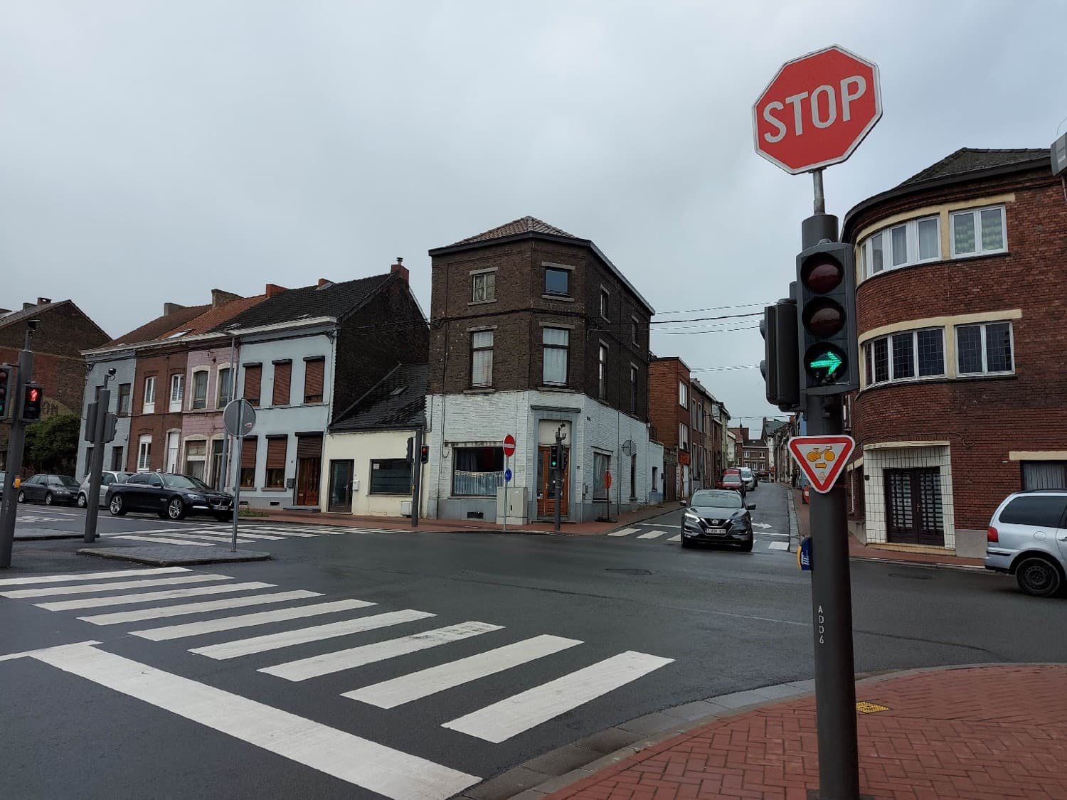 Absolument ! En tant qu'expert du code de la route belge, je vais analyser cette image en détail pour le contexte de votre question "Je souhaite tourner à droite à ce carrefour".
Description détaillée de la situation routière :
Type de situation routière :
Nous sommes face à un carrefour urbain complexe et réglementé, situé dans un quartier qui semble être résidentiel ou mixte (résidentiel avec quelques commerces au rez-de-chaussée). La chaussée est asphaltée et paraît légèrement humide, suggérant un temps couvert ou une pluie récente. L'intersection est encadrée par des bâtiments mitoyens en briques, typiques de l'architecture belge.
Éléments visibles (généraux) :
Chaussée et trottoirs : La chaussée est en asphalte. Les trottoirs sont pavés de briques rouges ou similaires, bien distincts de la route.
Bâtiments : Des maisons de ville à plusieurs étages, principalement en briques, bordent les rues du carrefour. Le bâtiment à l'angle droit présente une façade arrondie et de nombreuses fenêtres.
Ciel : Le ciel est gris et couvert, suggérant une journée nuageuse.
Marquages au sol : Un passage pour piétons (zébra) est clairement visible juste devant la position du conducteur, marquant l'entrée du carrefour.
Position et direction des véhicules :
Position du conducteur (implicite) : Le véhicule du conducteur (dont nous avons le point de vue) est arrêté ou sur le point de s'arrêter au début du carrefour, juste avant le passage pour piétons et la ligne d'arrêt (non visible mais implicite du fait du panneau STOP). Le conducteur se prépare à tourner à droite.
Véhicules à gauche : Deux voitures foncées (dont une BMW Série 5) sont stationnées sur le côté gauche de la rue, avant le carrefour.
Véhicule venant d'en face (rue de droite) : Une Nissan Qashqai noire est visible dans la rue de droite, face à nous, et semble s'engager dans le carrefour ou en sortir. Elle est en mouvement.
Véhicules garés à droite : Une voiture familiale de couleur gris clair est garée le long du trottoir, sur la rue où le conducteur souhaite tourner à droite, juste après le coin du bâtiment.
Véhicules au loin : Quelques autres véhicules sont visibles, plus loin dans la perspective de la rue d'en face.
Panneaux de signalisation et feux présents et leur signification (Code de la Route Belge) :
Sur le poteau de signalisation principal, à droite, on observe plusieurs éléments cruciaux :
Panneau STOP (B5) : C'est un panneau octogonal rouge avec le mot "STOP" en blanc. Sa présence indique une obligation d'arrêt absolu avant la ligne d'arrêt (ou, en son absence, à la hauteur du panneau) et de céder le passage à tous les autres usagers venant des autres branches du carrefour, y compris les piétons et cyclistes.
Feu de signalisation vertical (feu tricolore) :
Feu rouge principal (ou éteint/douteux pour la voie tout droit) : Les feux supérieurs et inférieurs du feu tricolore principal semblent être rouges ou éteints. Cela signifierait un arrêt obligatoire pour la direction "tout droit" ou la direction générale non-spécifiée par une flèche.
Feu directionnel vert avec flèche vers la droite : C'est une flèche verte lumineuse indiquant la direction à droite. En Belgique, ce feu permet de tourner à droite même si le feu principal est rouge. ATTENTION : La présence de cette flèche verte n'annule PAS les règles de priorité (notamment le STOP en l'occurrence) ni l'obligation de céder le passage aux usagers venant d'autres directions et aux piétons ou cyclistes qui traversent la voie sur laquelle vous allez vous engager.
Panneau "Cédez le passage" (B1) : Un panneau triangulaire blanc avec un bord rouge et un pictogramme de deux voitures. Il est situé sous le feu directionnel. Sa présence est quelque peu redondante avec le panneau STOP pour la priorité à l'entrée du carrefour, mais il renforce l'obligation de céder le passage après l'arrêt obligatoire et lors de l'exécution de la manœuvre de virage à droite, surtout si d'autres usagers sont concernés par la flèche verte.
Panneau pour piétons/cyclistes (à gauche) : Sur le poteau de gauche (devant les véhicules garés), un feu piéton/cycliste est visible et semble être rouge, indiquant que les piétons ou cyclistes venant de cette direction n'ont pas le droit de traverser.
Autres panneaux :
Sur le trottoir à l'extrême droite, après le carrefour, sur la rue dans laquelle on s'apprête à tourner, un panneau d'interdiction de stationner ou d'arrêt et de stationnement (type E1 ou E3) est visible, indiquant des restrictions pour le stationnement dans cette rue.
Tout autre élément pertinent pour comprendre la situation :
Passage pour piétons : La présence du zébra signifie que le conducteur doit être particulièrement vigilant aux piétons. Il doit s'arrêter avant le passage si des piétons s'y trouvent ou s'apprêtent à s'y engager, et de toute façon respecter son arrêt au panneau STOP avant le passage.
Coordination des signaux : Le carrefour est clairement régulé par une combinaison de feux de signalisation et de panneaux. La hiérarchie des signaux en Belgique est importante : le feu vert directionnel prime sur le panneau STOP dans le sens de la progression, MAIS l'arrêt imposé par le STOP (B5) reste une obligation avant de s'engager, et la flèche verte directionnelle implique de céder le passage aux autres usagers. Le panneau B1 (Cédez le passage) est une confirmation de cette obligation de prudence.
En résumé, pour le conducteur souhaitant tourner à droite, la situation exige :
Un arrêt obligatoire au panneau STOP (B5) avant le passage pour piétons et la ligne d'arrêt.
De céder le passage à tous les usagers venant des autres branches du carrefour et aux piétons et cyclistes traversant la chaussée sur laquelle il va s'engager, même si la flèche verte l'autorise à tourner à droite. La flèche verte ne donne pas une priorité absolue.
De s'assurer que le virage peut être effectué en toute sécurité.